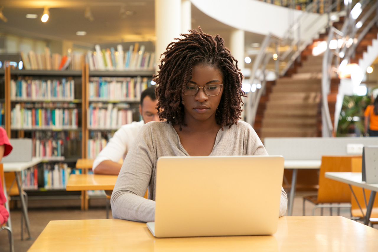 Mulher em uma biblioteca concentrada enquanto utiliza um notebook para fazer seu TCC. Um homem sentado na cadeira de trás estudando e, ao fundo, diversos livros em prateleiras