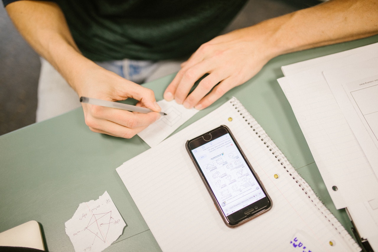 Foto de um homem fazendo uma anotação, enquanto utiliza o celular para estudar online. Na tela, estão os conteúdos referente à matéria e embaixo do celular, um caderno com exercícios.