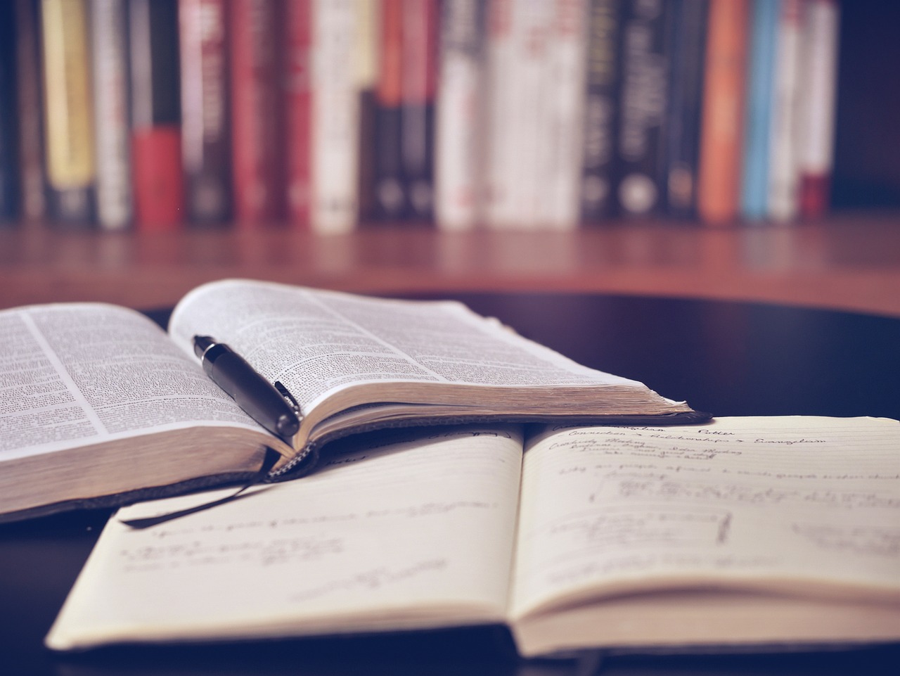 anotações em um caderno e um livro de estudos aberto em cima de uma mesa, com mais livros nas prateleiras ao fundo