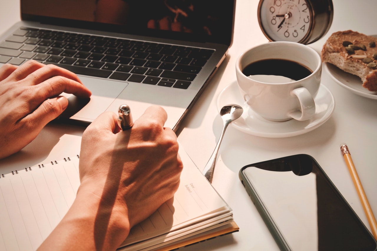 Homem utilizando o Método Pomodoro para realizar suas tarefas. A imagem mostra apenas suas mãos sobre uma mesa de madeira, enquanto ele faz anotações em um caderno e utiliza um notebook. Na mesa também estão um relógio de ponteiro, uma xícara de café, um celular e um prato com sanduíche.