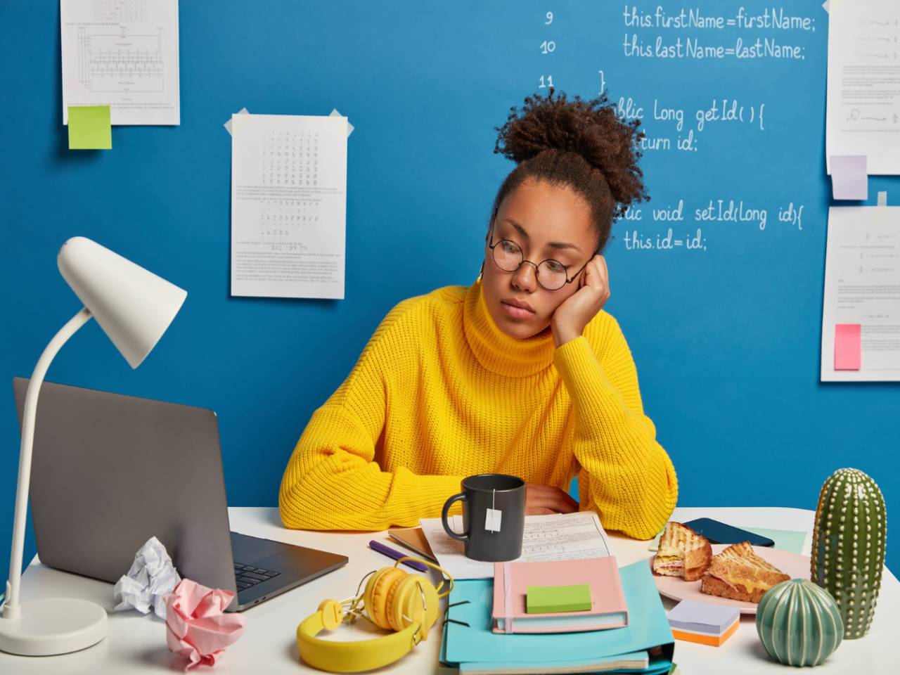 Estudante sentada em uma cadeira com expressão de tédio e o rosto apoiado em uma das mãos, buscando uma motivação para estudar. À sua frente, uma mesa com livros, notebook e outros materiais de estudos
