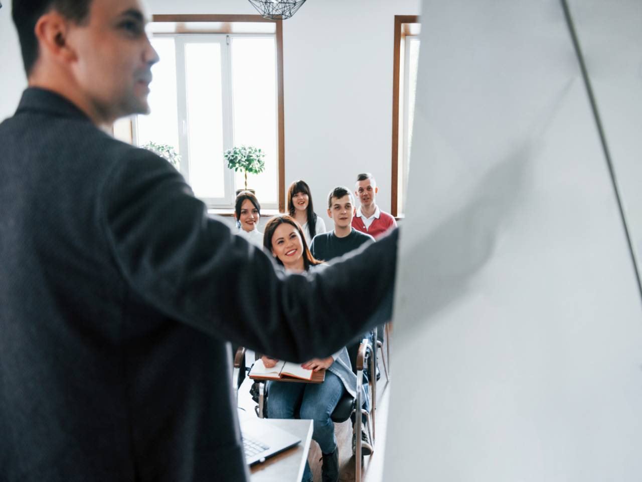 um homem de lado apontando para um quadro branco. fazendo uma apresentação de trabalho. Ao fundo, alguns alunos de frente para ele, prestando atenção.
