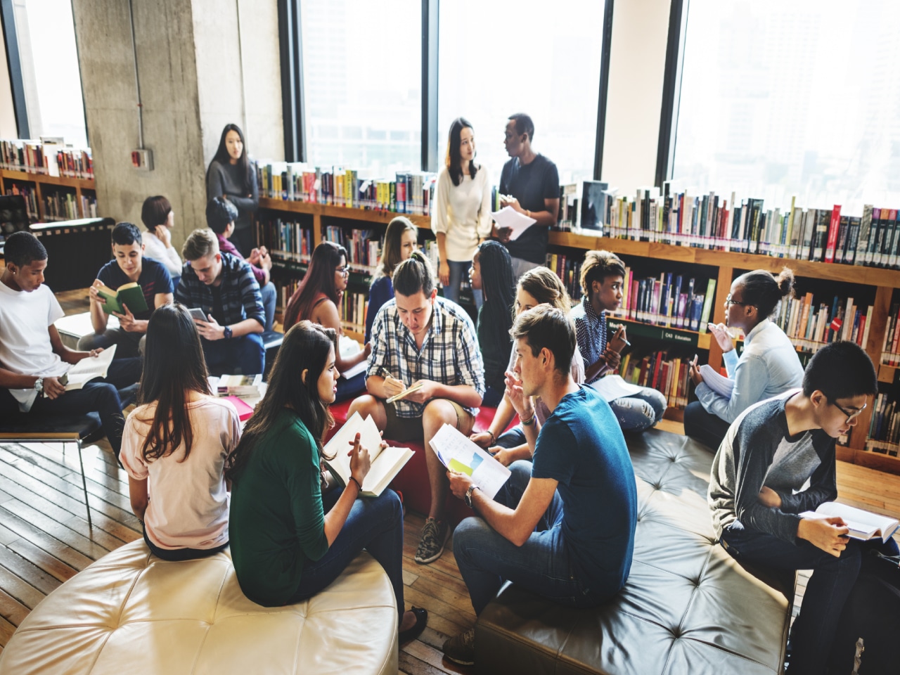 Vários alunos que fazem parte de um grupo de estudo, reunidos em uma biblioteca. Alguns estão de pé, enquanto outros estão sentados debatendo e lendo livros sobre a matéria.