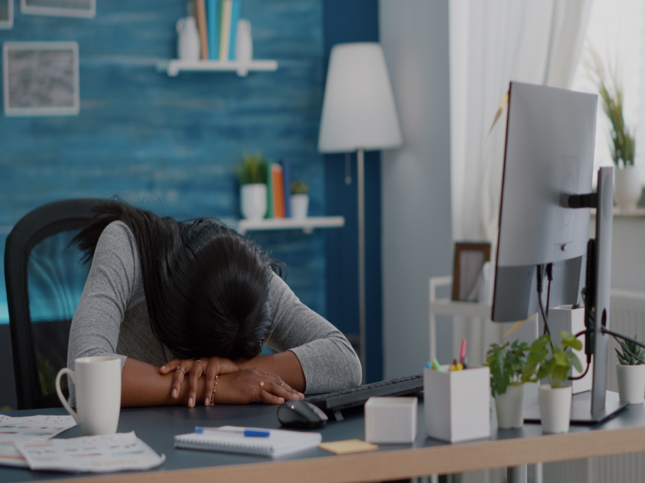 Como regular o sono: estudante em seu quarto debruçada sobre sua mesa, dormindo durante o período de estudo. Na mesa, estão seu computador, alguns enfeites e papéis, e uma xícara.