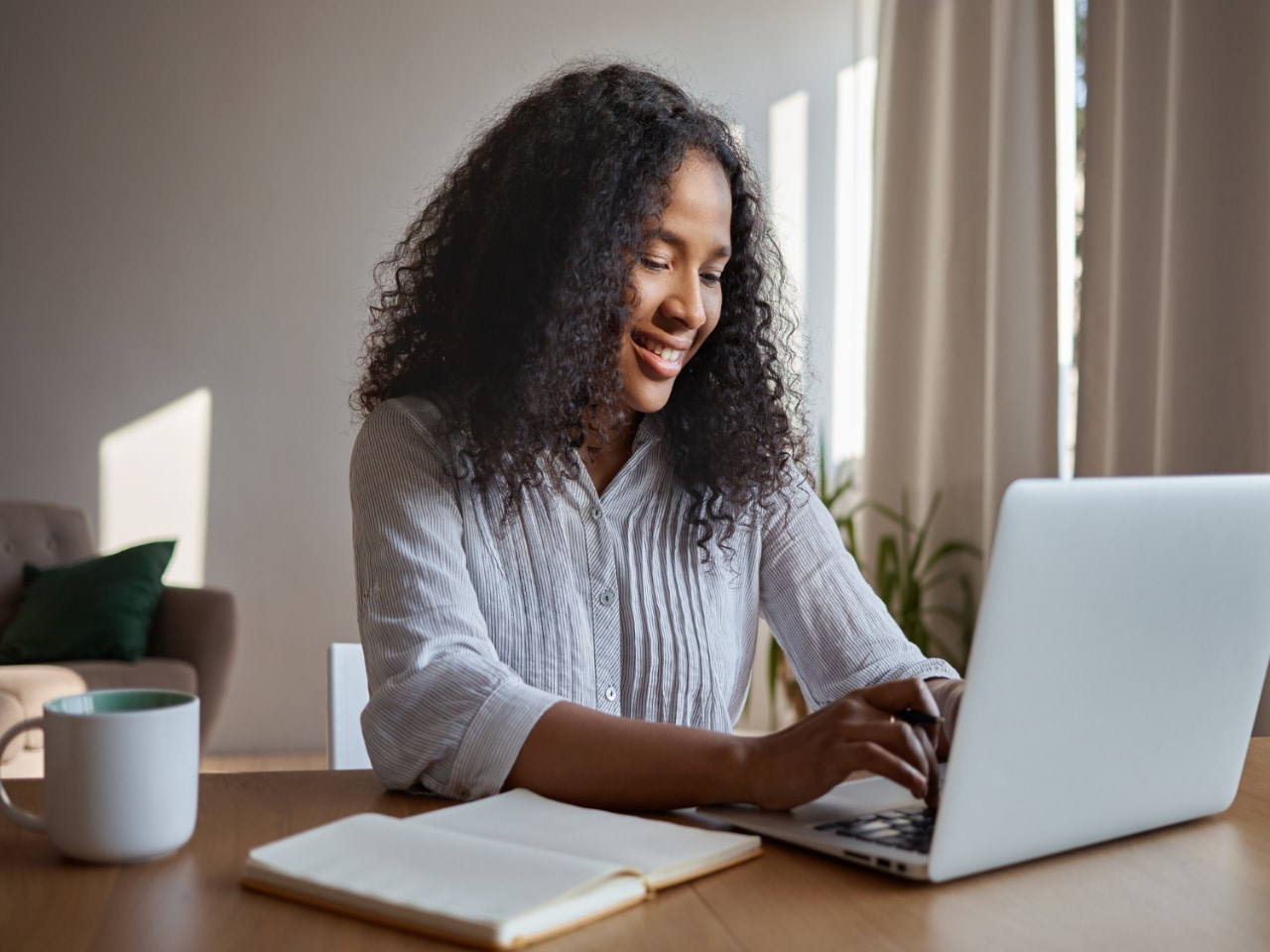Estudar em casa: uma jovem sorridente sentada em sua mesa de estudos, mexendo em um notebook. Ao seu lado, um caderno aberto e uma caneca com alguma bebida.