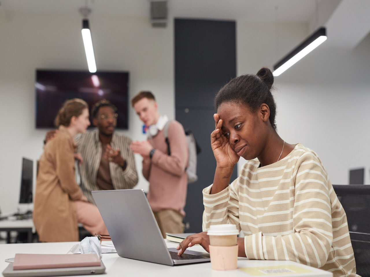 O que é ergofobia: mulher sentada em sua cadeira de escritório com um notebook à sua frente. Ela está com uma das mãos na cabeça e com uma expressão de desconforto, enquanto três colegas de trabalho estão ao fundo conversando e olhando para ela.