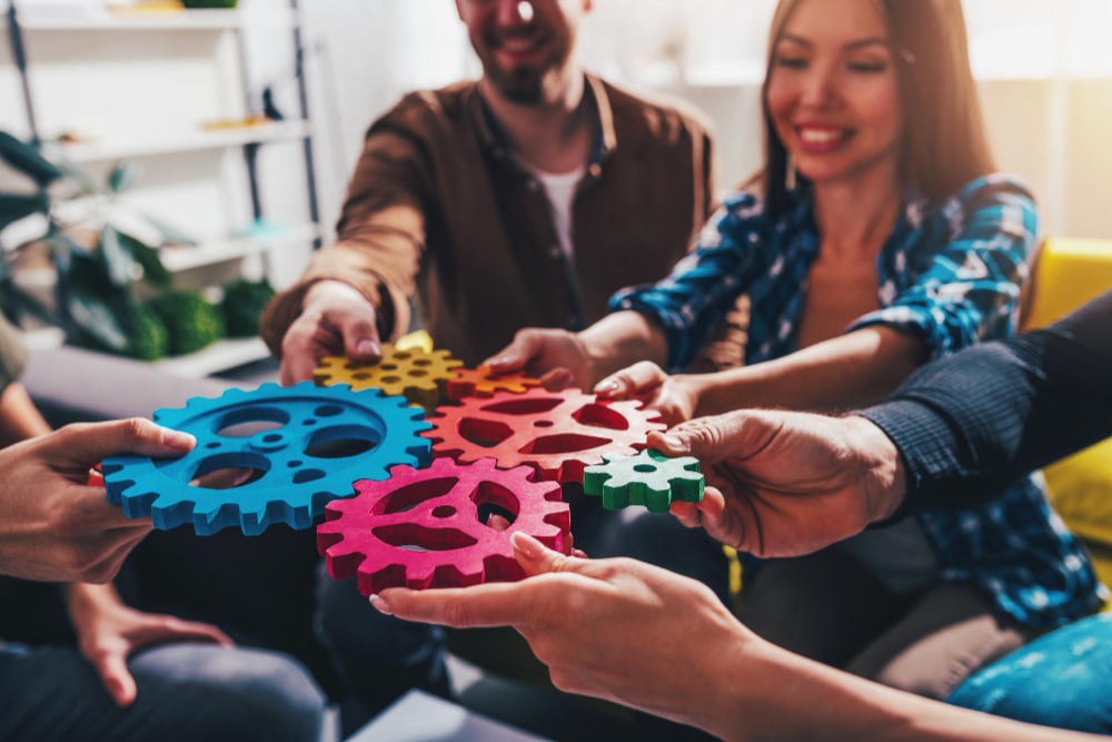 Cinco funcionários sorridentes em um escritório, sentados juntos em um sofá fortalecendo suas habilidades interpessoais. Cada um segura uma engrenagem para que elas se encaixem corretamente, representando o trabalho em equipe.